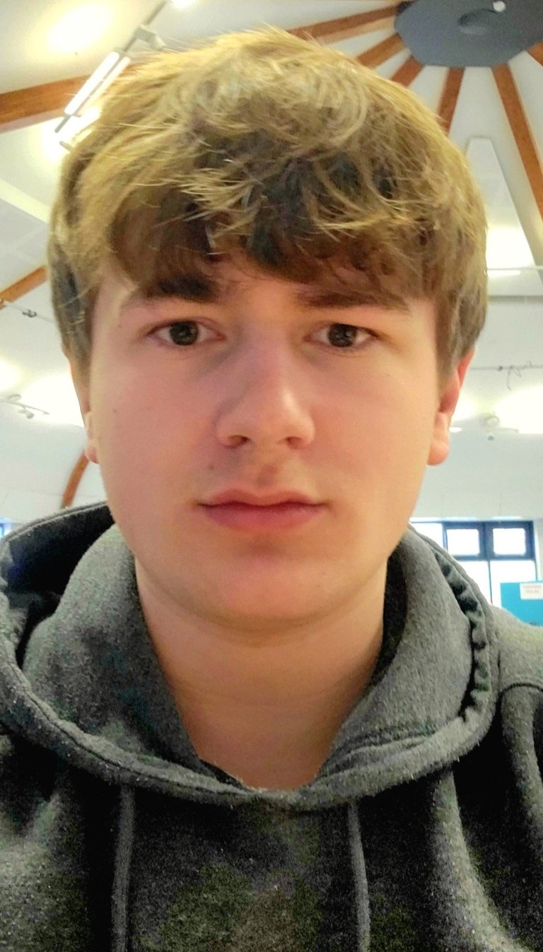 Close-up portrait of a young person with blonde hair and bangs wearing a gray hoodie, photographed indoors under ceiling lights